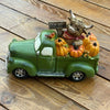 A decorative green vintage pickup truck figurine loaded with orange pumpkins, a brown highland cow, and a small 'Welcome' sign, displayed on a white faux-fur surface with a wooden bowl in the background.  Available at Quilted Cabin in Airdrie