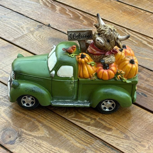A decorative green vintage pickup truck figurine loaded with orange pumpkins, a brown highland cow, and a small 'Welcome' sign, displayed on a white faux-fur surface with a wooden bowl in the background.  Available at Quilted Cabin in Airdrie