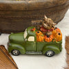 A decorative green vintage pickup truck figurine loaded with orange pumpkins, a brown highland cow, and a small 'Welcome' sign, displayed on a white faux-fur surface with a wooden bowl in the background.  Available at Quilted Cabin in Airdrie