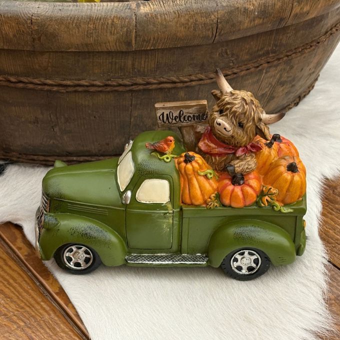 A decorative green vintage pickup truck figurine loaded with orange pumpkins, a brown highland cow, and a small 'Welcome' sign, displayed on a white faux-fur surface with a wooden bowl in the background.  Available at Quilted Cabin in Airdrie