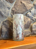 A cylindrical glass candle holder or vase with a frosted off-white background and a printed design of red cardinals on branches with red berries, set on a wooden surface against a stone wall.  Available at Quilted Cabin in Airdrie
