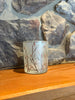 A small frosted glass candle holder featuring a delicate branch and cherry blossom pattern in brown and pink, resting on a wooden surface against a rustic stone wall background.  Available at Quilted Cabin in Airdrie