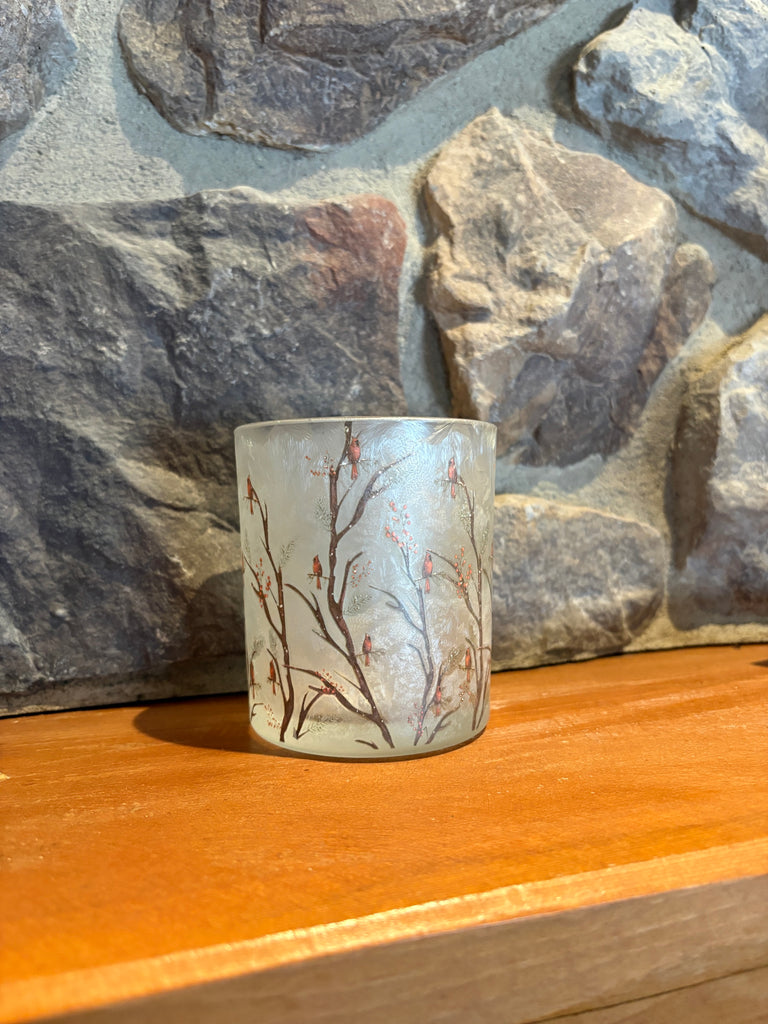 A small frosted glass candle holder featuring a delicate branch and cherry blossom pattern in brown and pink, resting on a wooden surface against a rustic stone wall background.  Available at Quilted Cabin in Airdrie