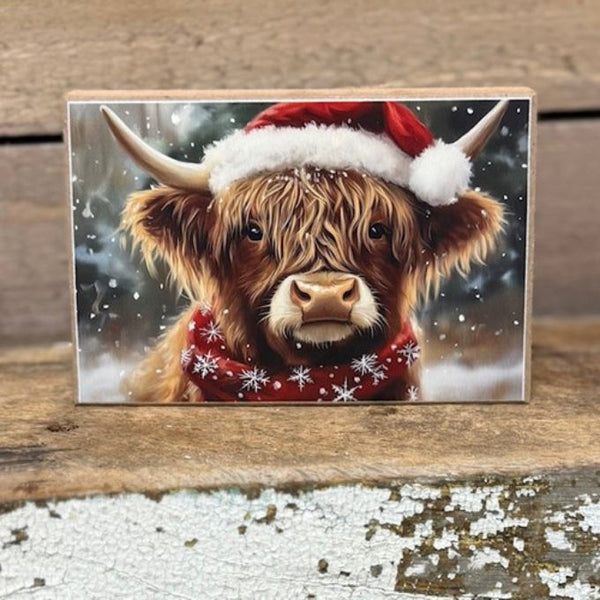 Wooden Block with an image of a highland cow wearing a Santa hat and scarf on a wooden surface. Winter Highland Art Block available at Quilted Cabin Home Decor.