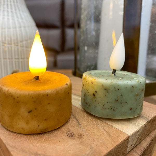 Two lit candles on a wooden surface with blurred background. LED Tea Lights - Two Colours available at Quilted Cabin Home Decor.