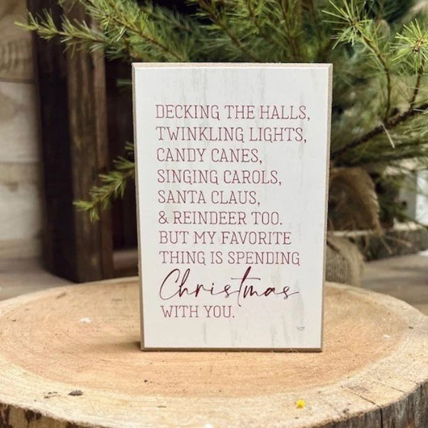 A rectangular white wooden sign with a rustic finish, featuring a printed Christmas quote in dark red lettering. The sign is displayed on a rustic wood slice with pine branches in the background.  Available at Quilted Cabin in Airdrie