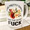 A white ceramic coffee mug. It is imprinted on both sides with a pretty display of flowers in the center and around the flowers the words, I like pretty things and the word F-U-C-K.