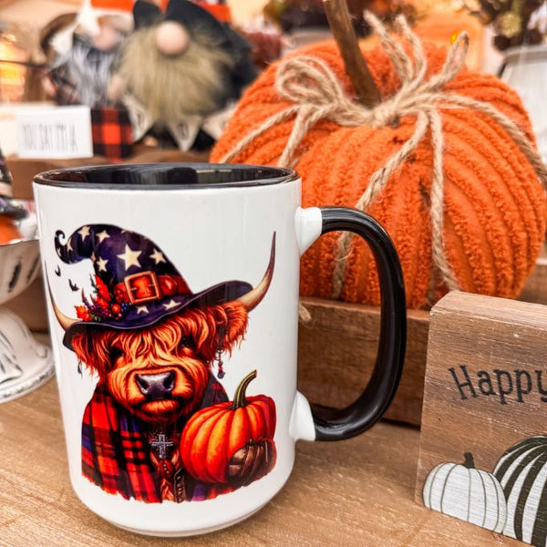 Mug with a Highland cow wearing a witch hat and holding a pumpkin, placed on a wooden surface with pumpkins and a sign in the background. The Halloween Highland Cow Mug is available at Quilted Cabin  Home Decor.