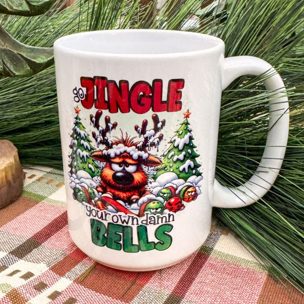 White mug with Christmas-themed design featuring a reindeer and text, placed on a checkered tablecloth with greenery in the background. Jingle Your Own Bells Mug available at Quilted Cabin Home Decor.