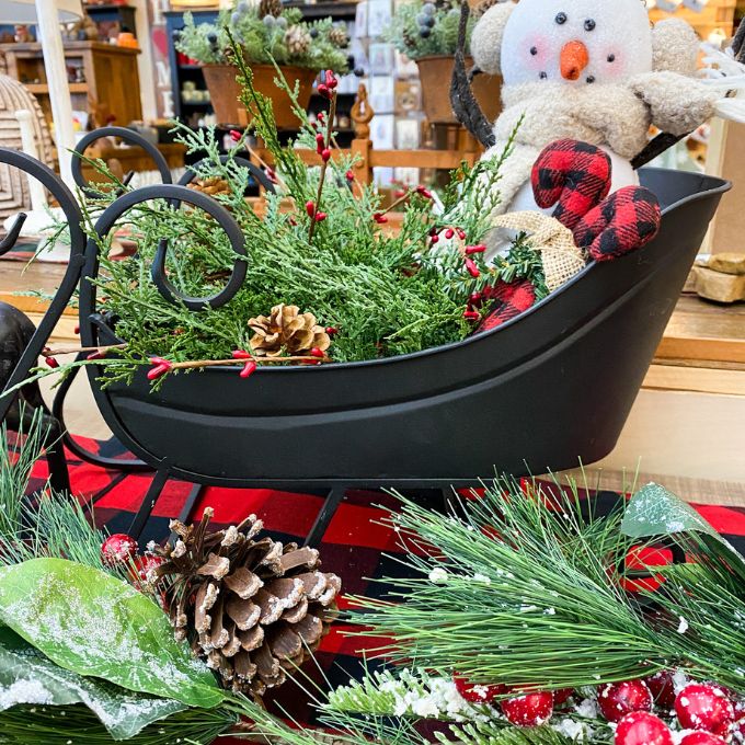 Decorative arrangement with greenery, pinecones, and a snowman figure on a checkered tablecloth. Berry and Cedar Dripping Floral Orbs - Two Styles available at Quilted Cabin Home Decor.
