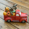 A decorative green vintage pickup truck figurine loaded with orange pumpkins, a brown highland cow, and a small 'Welcome' sign, displayed on a white faux-fur surface with a wooden bowl in the background.  Available at Quilted Cabin in Airdrie