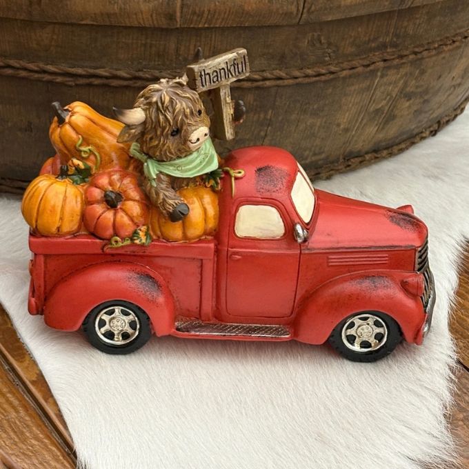 A decorative green vintage pickup truck figurine loaded with orange pumpkins, a brown highland cow, and a small 'Welcome' sign, displayed on a white faux-fur surface with a wooden bowl in the background.  Available at Quilted Cabin in Airdrie