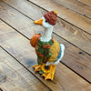 A decorative goose figurine wearing a brown knit beanie, green and brown knit sweater, and matching scarf, holding a small orange pumpkin, set on a wooden plank surface.  Available at Quilted Cabin in Airdrie