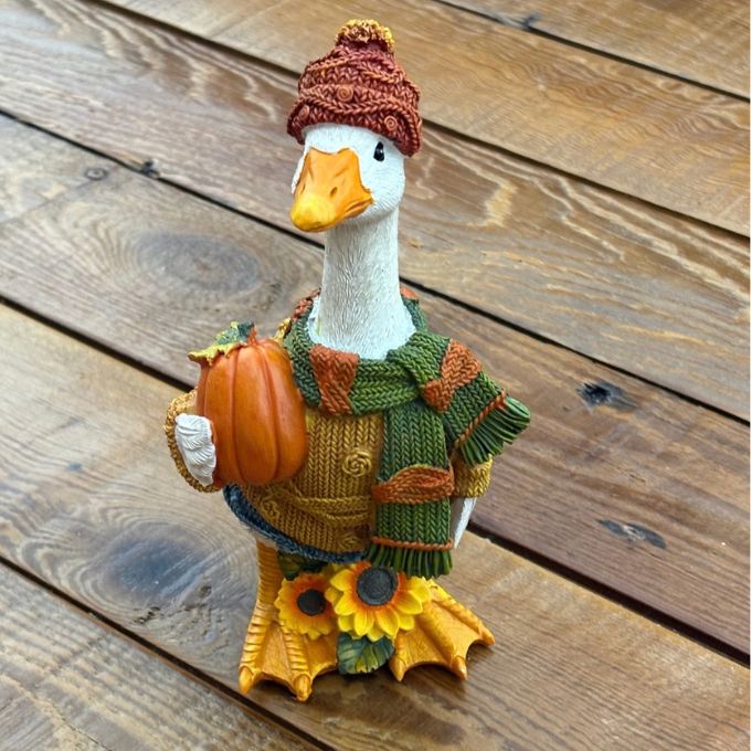 A decorative goose figurine wearing a brown knit beanie, green and brown knit sweater, and matching scarf, holding a small orange pumpkin, set on a wooden plank surface.  Available at Quilted Cabin in Airdrie