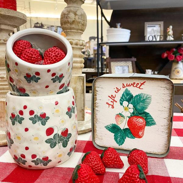 Decorative strawberry-themed containers with a small sign on a red checkered tablecloth. Strawberry Block Sign available at Quilted Cabin Home Decor.