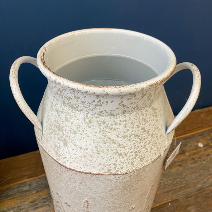 White ceramic vase with handles on a wooden surface against a blue background. Embossed Metal Milk Cans - Two Colours available at Quilted Cabin Home Decor.