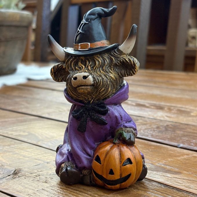 A small decorative figurine of a shaggy Highland cow dressed as a witch, sitting on a wooden surface. The figurine features a black cape with red lining, a black bow on its head, and a metallic gold-brown textured coat. Available at Quilted Cabin in Airdrie