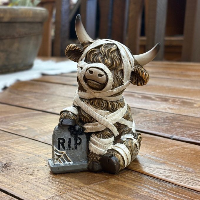 A small Highland cow figurine wrapped in beige bandages and holding a gray tombstone with 'R.I.P" available at Quilted Cabin in Airdrie