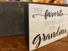 A small wooden block sign that has black sides, and grey and white text on the front. The text says My favorite people call me grandma. The text is on distressed white/grey background. It is 2 3/4" tall and 4 1/2" wide. It is 3/4" deep and is a sign that will sit on the shelf.