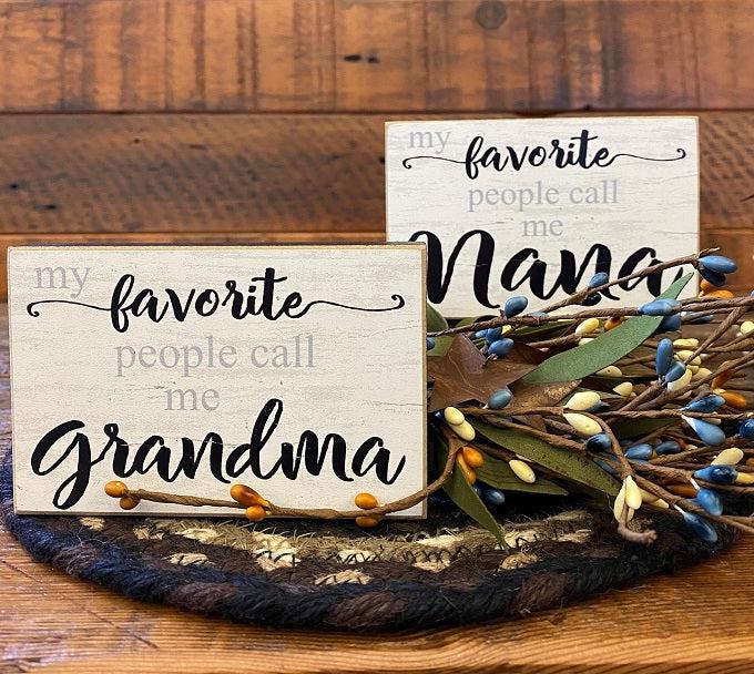 Two mini block signs and The edges are black and the writing is gray and black.  one sign says my favorite people call me grandma and the other says my favorite people call me Nana 