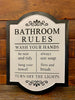 A white painted metal sign, painted with black trim and the words are all in Black.  It is a Bathroom rules sign with the following rules - wash your hands, be neat and tidy, hang your towel, always use soap, floss and brush and turn off the lights. 
