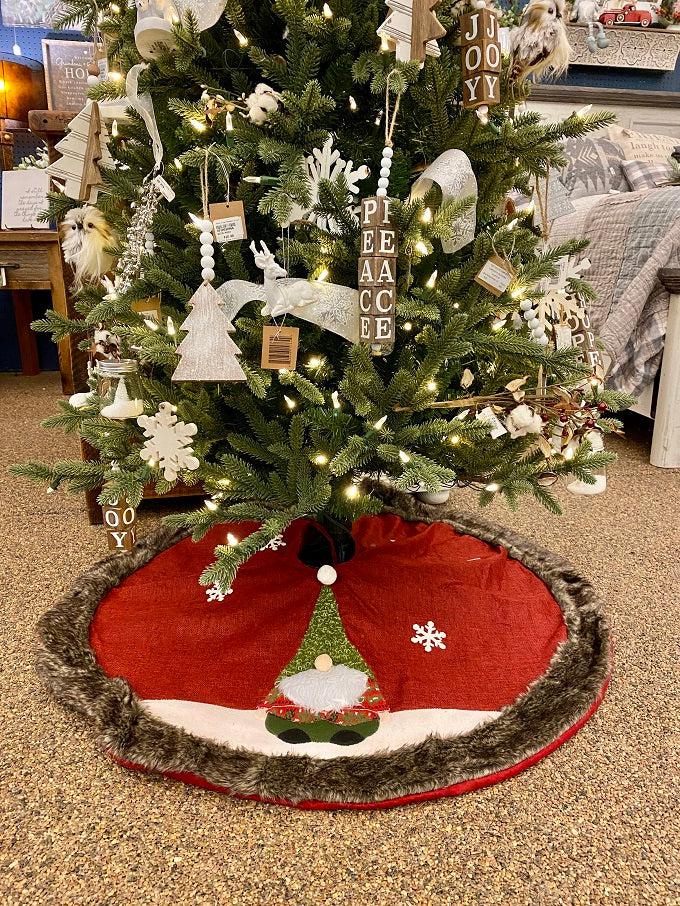 The Kringle Gnome tree skirt in red is shown. It has a festive gnome appliqued on it along with snowflakes. It is trimmed in faux fur.