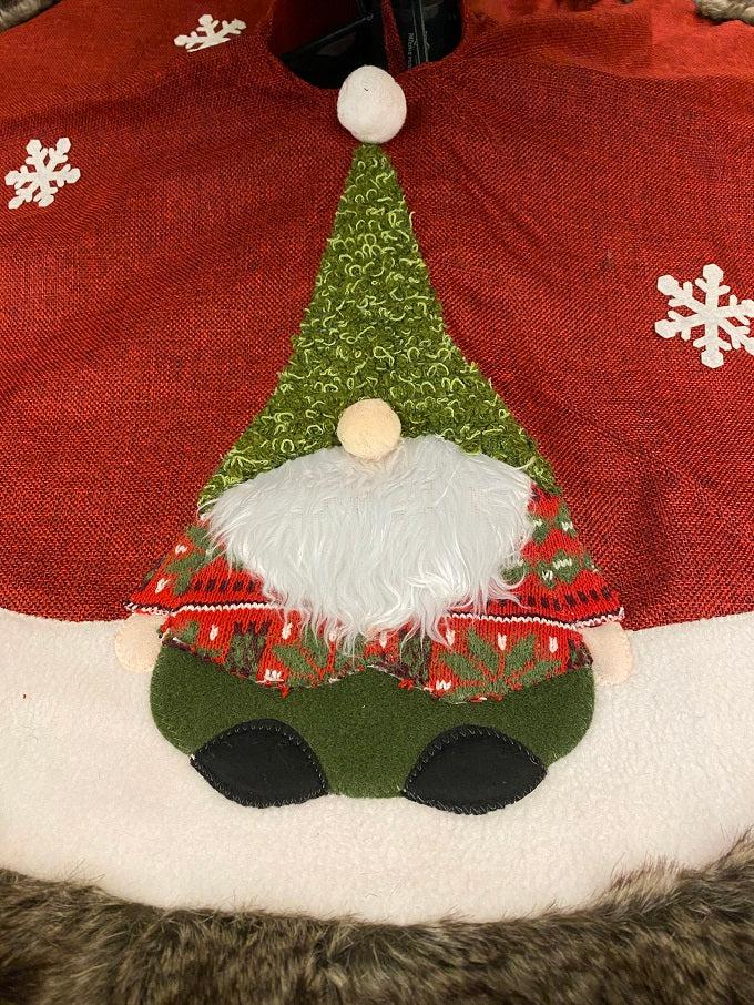 The Kringle Gnome tree skirt in red is shown. It has a festive gnome appliqued on it along with snowflakes. It is trimmed in faux fur.