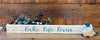  An 18" long white, 3/4" wide and 1" tall shelf sitting sign. It has a planked looked background and on the front is written in blue script font Lake Life Livin.