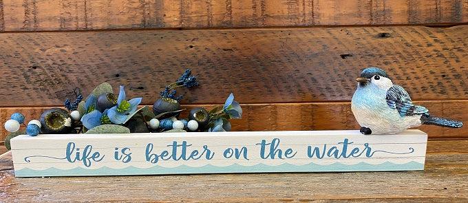 An 18" long white, 3/4" wide and 1" tall shelf sitting sign. It has a planked looked background and on the front is written in script life is better on the water. There is painted waves along the whole length of the sign on the front.