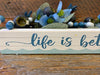 A close up photo of the Life is better on the water shelf sitting sign. It shows the blue script writing, and the painted waves running along the bottom of the sign.