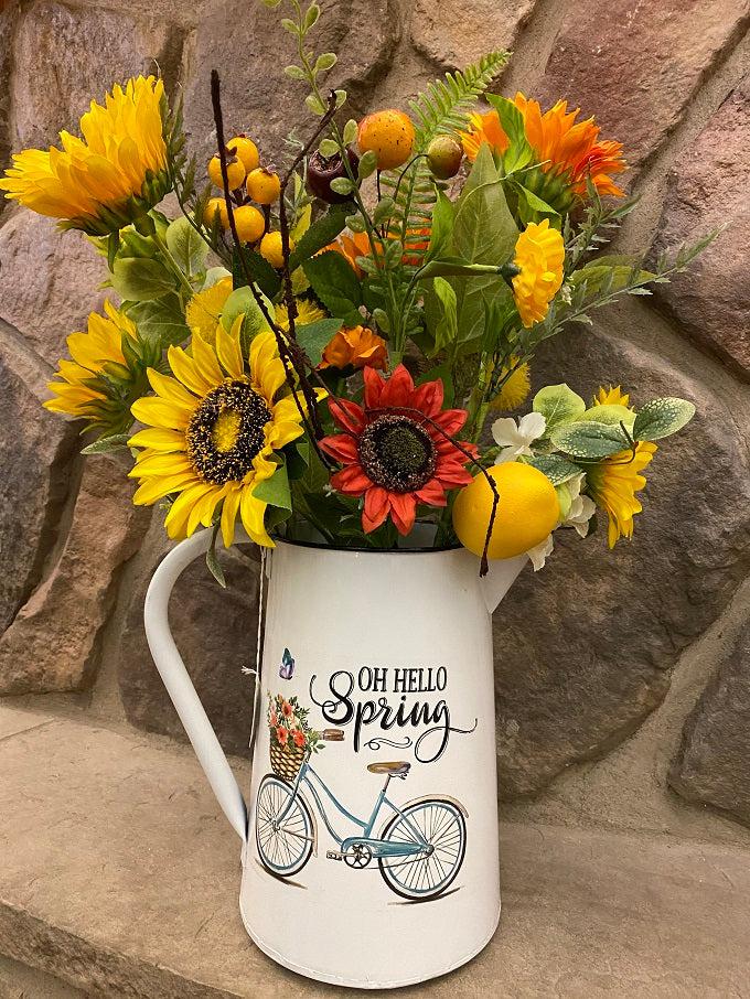 A white enamel pitcher is filled with beautiful sunflowers. The pitcher is white enamel, and has a white handle, and black painted trim around the top and spout. The picture on the side is of a blue vintage bike and says Oh Hello Spring.