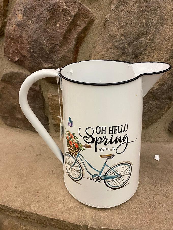 A close up of a white enamel pitcher. The picture on the side is of a blue vintage bike with a basked full of pink toned flowers and says Oh Hello Spring.