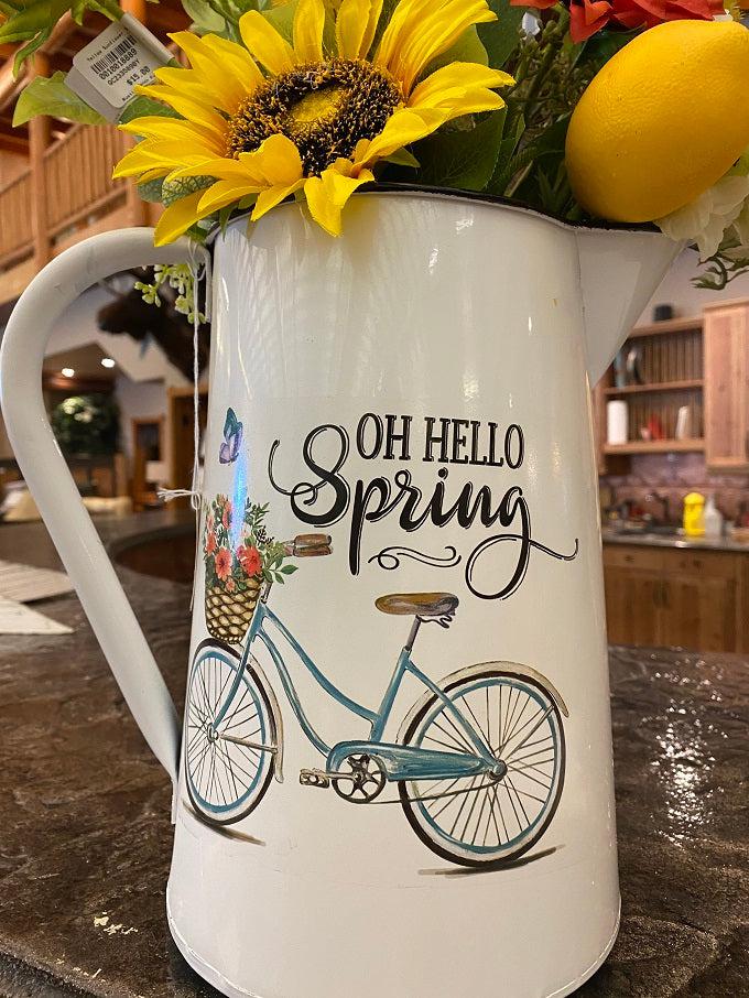 A close up of a white enamel pitcher. The picture on the side is of a blue vintage bike with a basked full of pink toned flowers and says Oh Hello Spring.