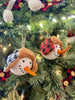 Two Holiday snowmen ornaments are shown. These ornaments are the head of the snowman with eyes made to look like coal and a carrot-like nose. One has a black and white plaid hat and the other has a red and black plaid hat.