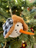 The Black and White Holiday Snowman Ornament is the head of a snowman with a black and white cap with fuzzy ear flaps.  He has two eyes made to look like coal and a carrot-like nose.