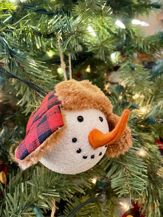 The Red and Black Holiday Snowman Ornament is the head of a snowman with a red and black cap with fuzzy ear flaps.  He has two eyes made to look like coal and a carrot-like nose.