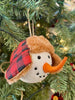 The Red and Black Holiday Snowman Ornament is the head of a snowman with a red and black cap with fuzzy ear flaps.  He has two eyes made to look like coal and a carrot-like nose.