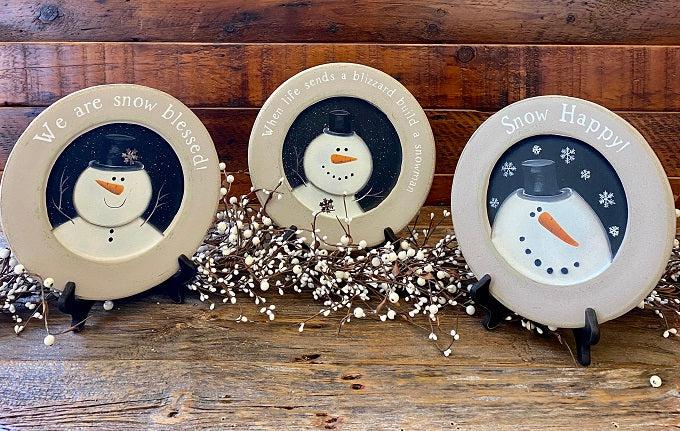 Three wooden snowman plates for your farmhouse holiday decorating. There are three styles to choose from: Snow Happy, We are Snow Blessed and When life sends a blizzard build a snowman. The edges of the plates are gray and the centre portion of the plate handpainted with the white snowman on a black background. The Snow Blessed and the Blizzard plate each have a tiny metal snowflake on them as well. 