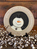 A farmhouse style wooden plate that says When Life sends a blizzard build a snowman. The plate has gray on the plate rime and the snowman is painted white and is on a black background.