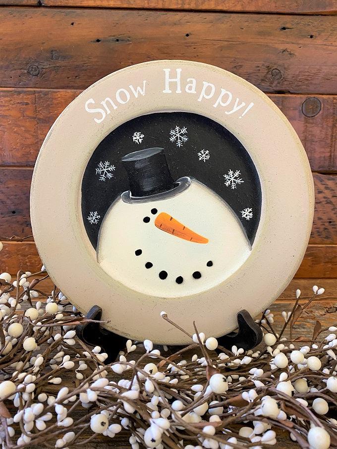 A farmhouse style wooden plate that says Snow Happy. The plate has gray on the plate rime and the snowman is painted white and is on a black background.