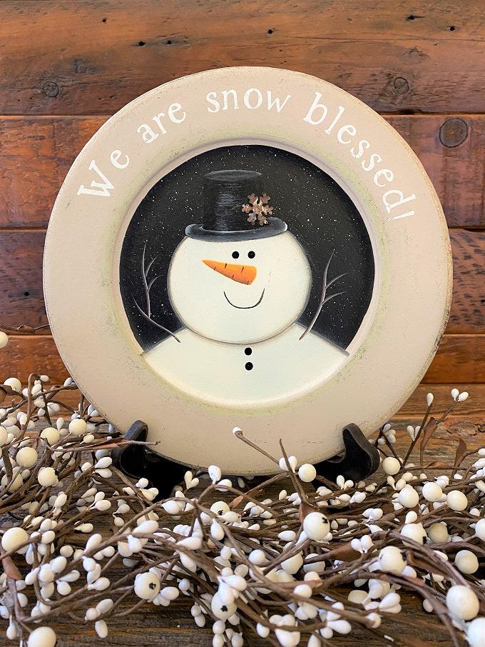 A farmhouse style wooden plate that says We are Snow Blessed. The plate has gray on the plate rime and the snowman is painted white and is on a black background.