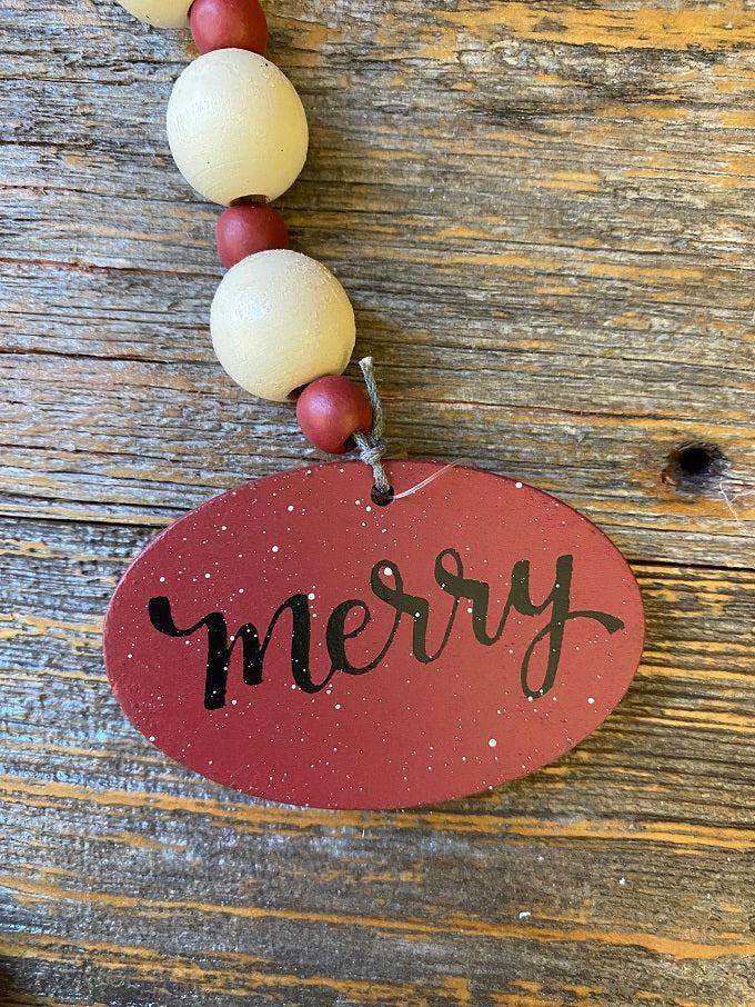 The merry wooden beaded garland is apporximately 19" long and made of white wooden beads alternately with smaller red wooden beads. At one end of the garland is a red wooden medallion with the word Merry painted on it in black lettering. At the other end is holly-like green leaf.  This photo is a close up of the merry medallion.