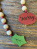 The merry wooden beaded garland is apporximately 19" long and made of white wooden beads alternately with smaller red wooden beads. At one end of the garland is a red wooden medallion with the word Merry painted on it in black lettering. At the other end is holly-like green leaf. 