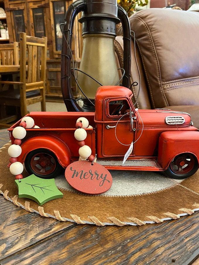 The merry wooden beaded garland is shown here in the back of a red metal truck. The garland is apporximately 19" long and made of white wooden beads alternately with smaller red wooden beads. At one end of the garland is a red wooden medallion with the word Merry painted on it in black lettering. At the other end is holly-like green leaf. 