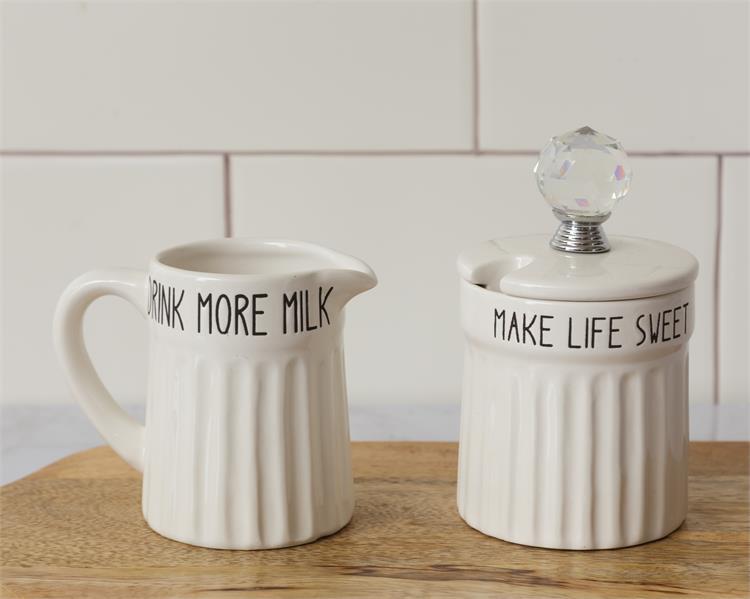White farmhouse inspired cream and sugar set. The body of both pieces are ridged and the rim around the top of each has black lettering. The milk jug say Drink more milk and the sugar bow says make life sweeter. The lid on the sugar bowl has a glass knob.