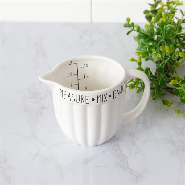 A white ceramic measuring cup that is a two cup size. The measure marks are in black on the inside of the cup. Around the outside rim of the cup are the words measure, mix and enjoy in black lettering. The cup has a handle and spout for easy pouring. 