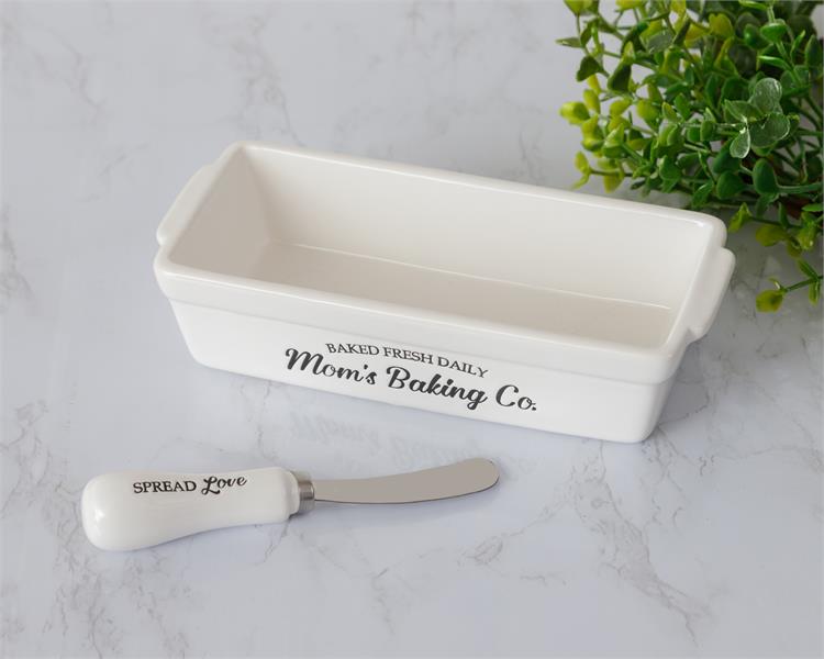 White rectangle shaped butter dish is shown with a ceramic handled server as well. The side of the white dish has black imprinted words that say Mom's Baking Co., baked fresh daily. The spreader says spread love.