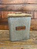 Galvanized flour canister made of corrugated metal. The canister is square with rounded corners or edges, The name plate on the side is copper. The lid is made of wood with a rectangle shaped handle.