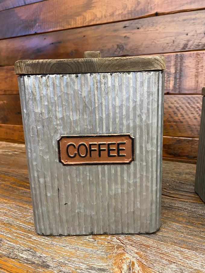 Galvanized coffee canister made of corrugated metal. The canister is square with rounded corners or edges, The name plate on the side is copper. The lid is made of wood with a rectangle shaped handle.