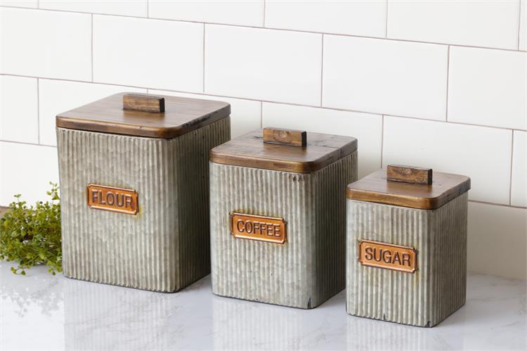 Set of three farmhouse style galvanized corrugated canisters. One says Flour, one says coffee and one says sugar. The name plates are copper. The lids are wooden and have wooded rectangle handles.. The tins are square with rounded corners, 